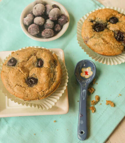 overhead shot of blueberry muffins on a turquoise linen baked in mint green paper liners with a bowl of fresh blueberries